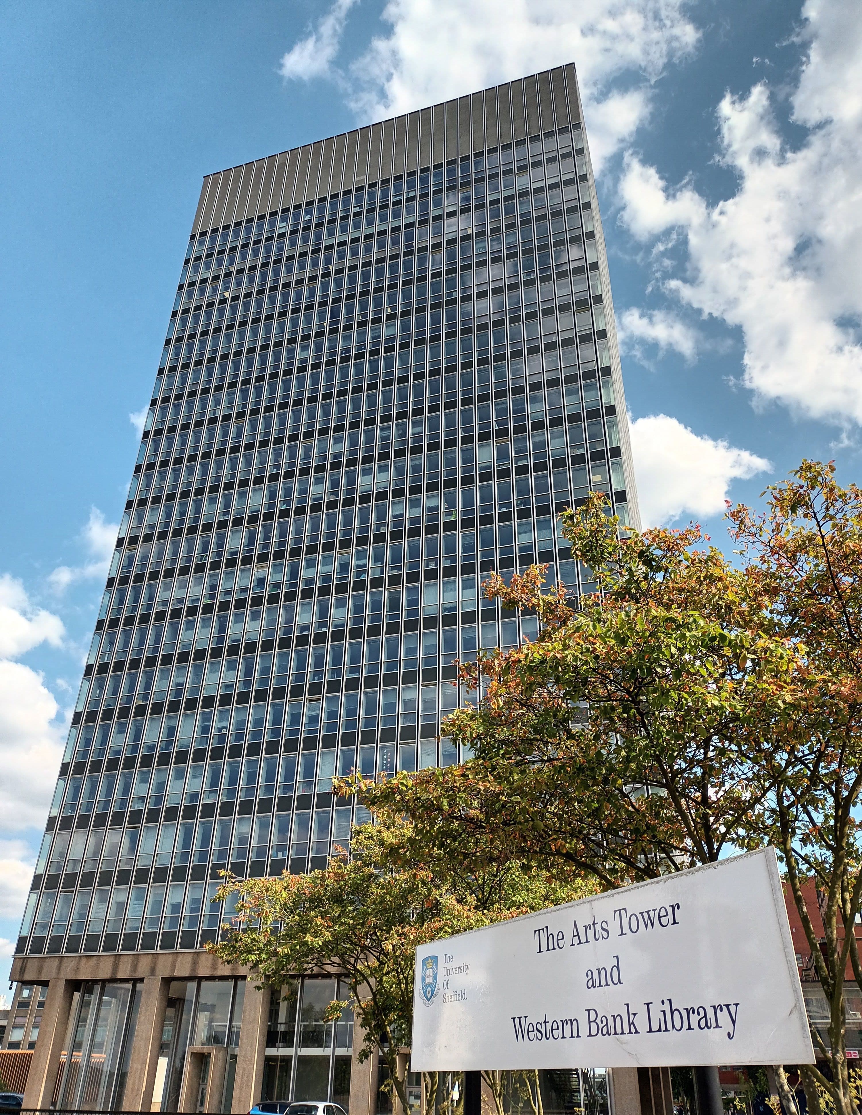 A tall, modern library tower building under a blue sky, with a sign in the foreground.