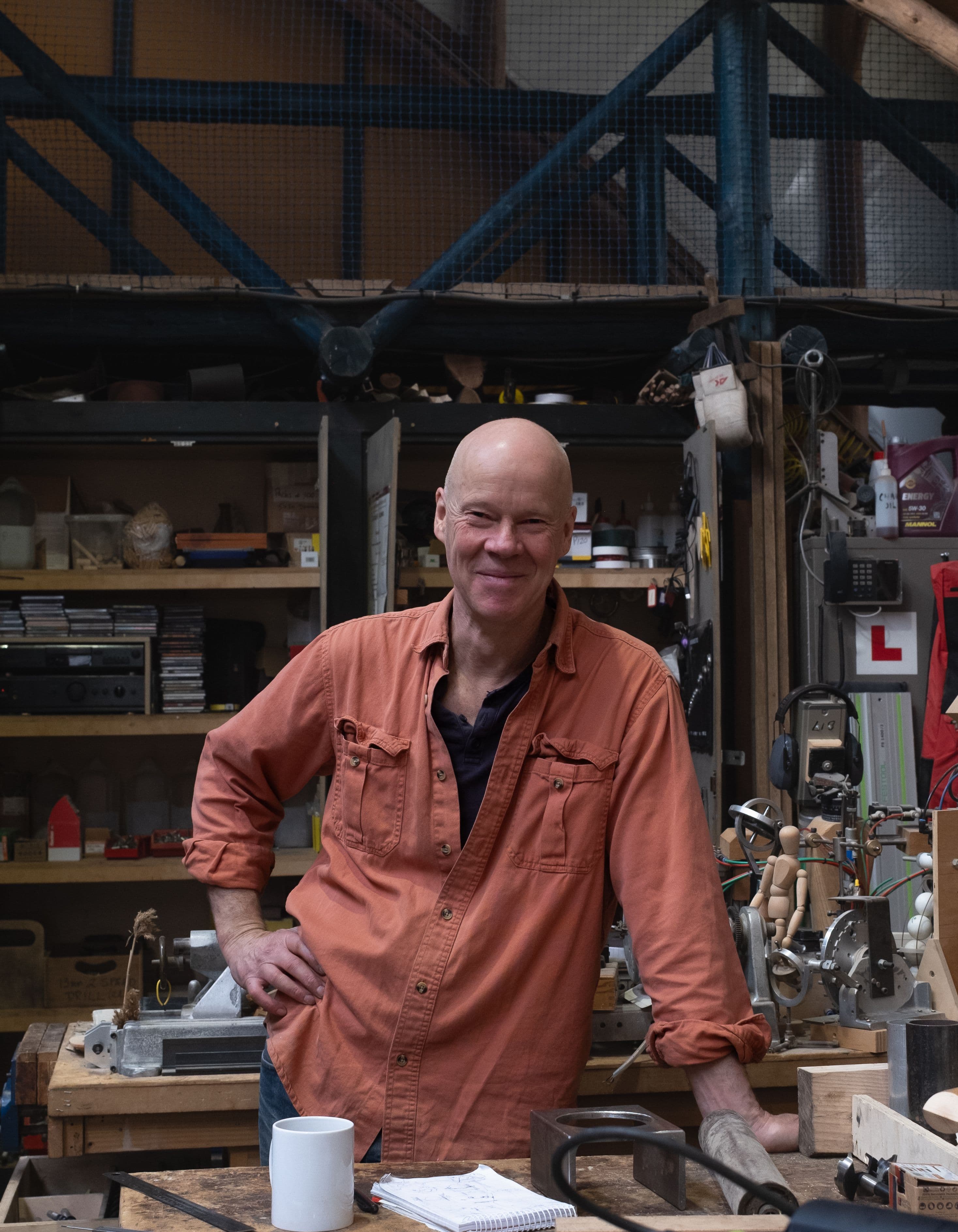 A white skinned man in an orange shirt stands smiling in a workshop, surrounded by tools.