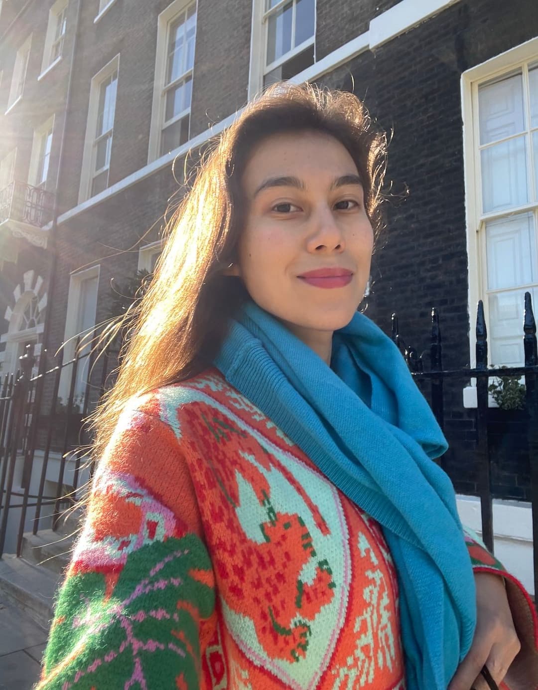 Woman wearing a colorful dragon-patterned sweater and a blue scarf, smiling outdoors with a building in the background.