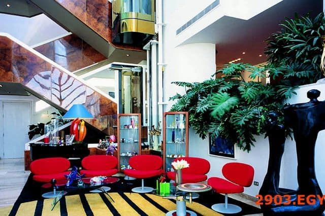 Modern hotel lobby with red lounge chairs, a grand staircase, and a glass elevator.