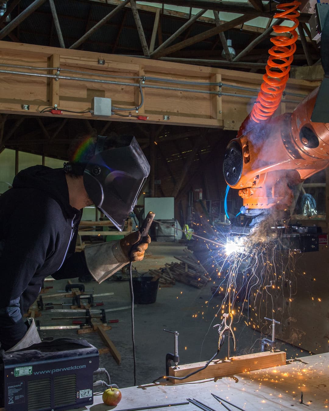 A man wearing a welding mask in a workshop welding onto an orange kuka robot arm