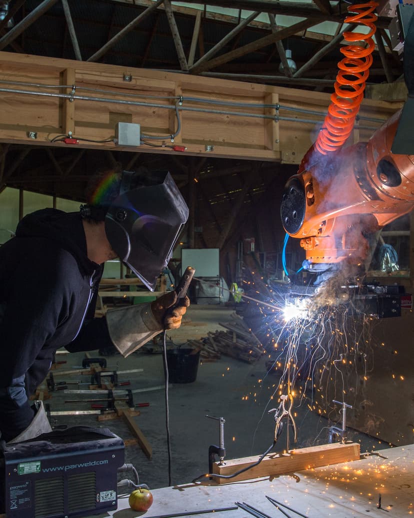 A man wearing a welding mask in a workshop welding onto an orange kuka robot arm