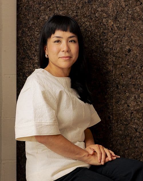 A woman with dark hair and bangs wears a white top and looks directly at the camera.