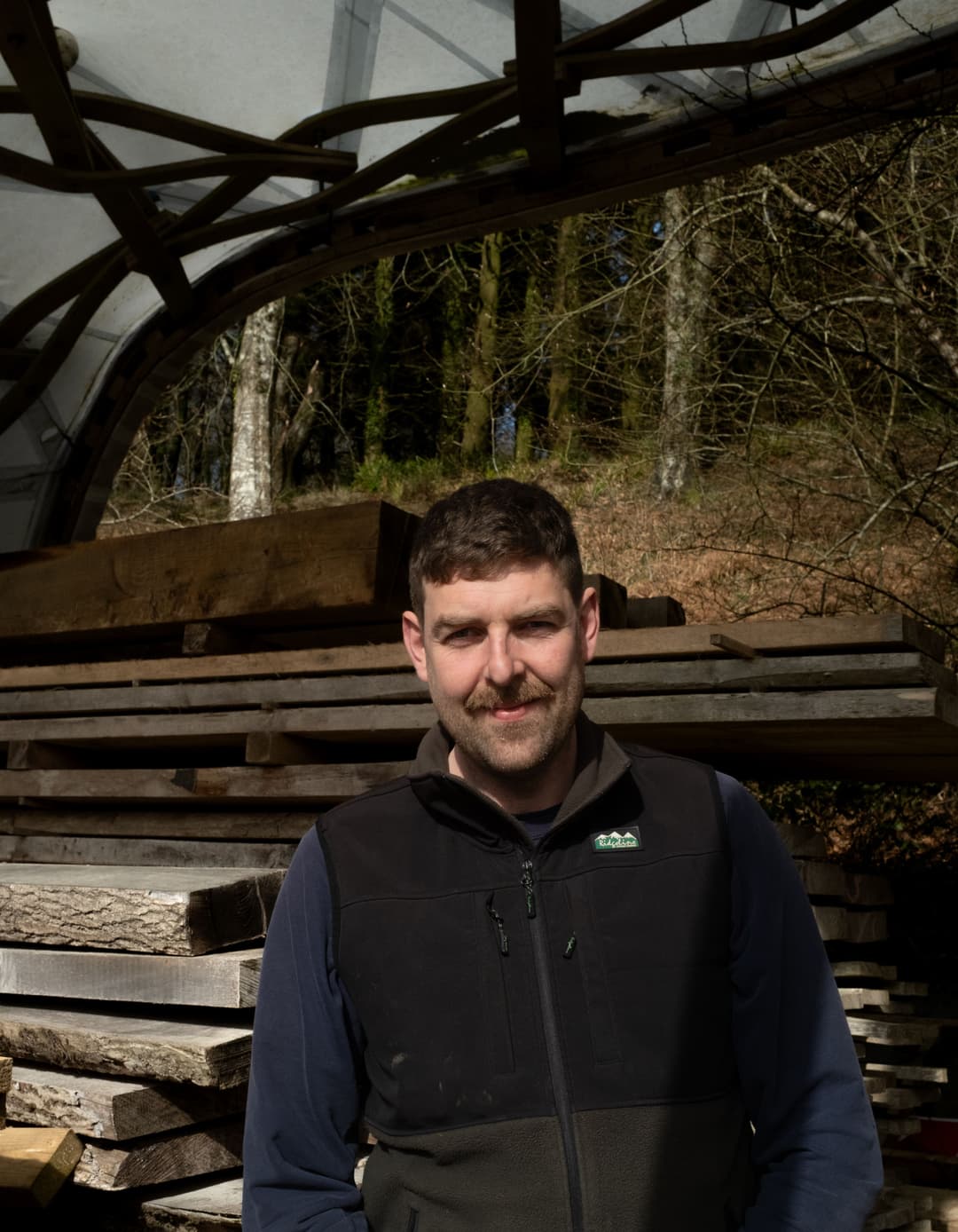 A white skinned man with short brown hair and a beard stands in a woodshed next to a pile of sawn timber.