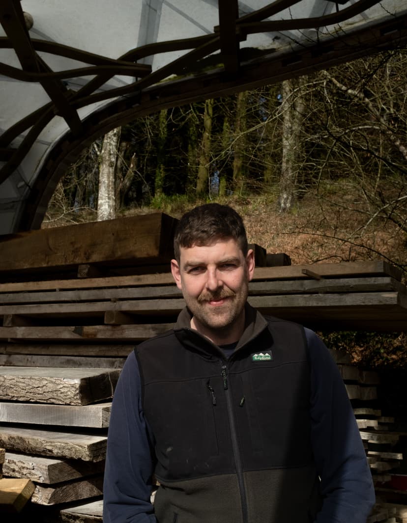 A white skinned man with short brown hair and a beard stands in a woodshed next to a pile of sawn timber.