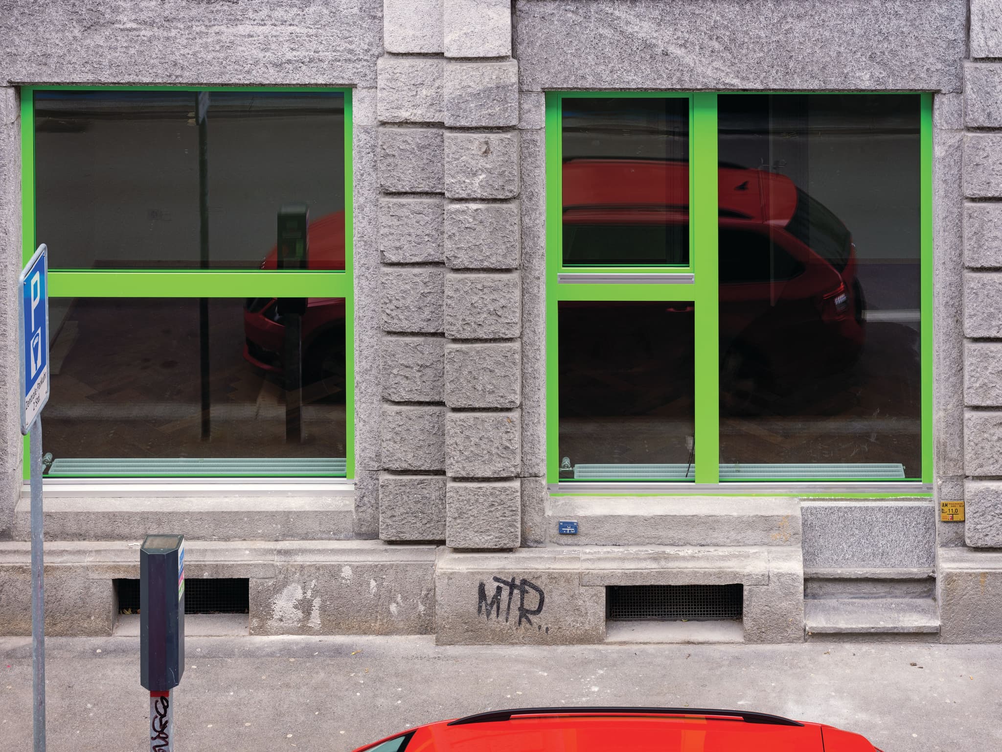 A stone building facade with bright green window frames. The top of a bright red car is visible in front of the building.