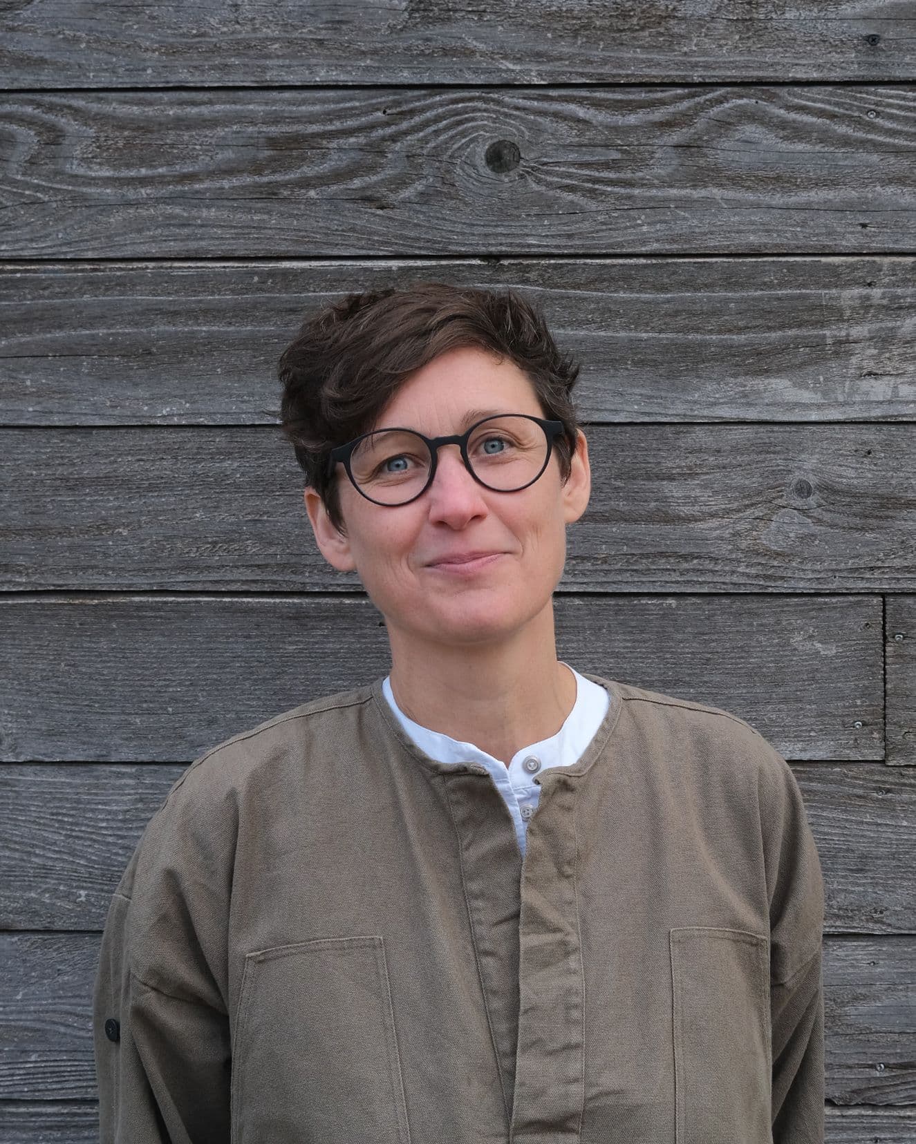 A white skinned woman with short brown hair and glasses stands against a timber wall.