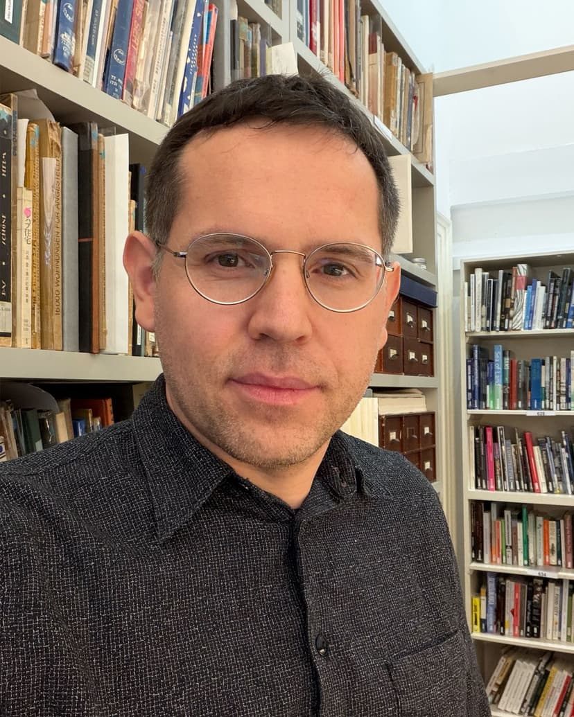 A man with short brown hair and glasses wears a dark collared shirt in front of a bookshelf.