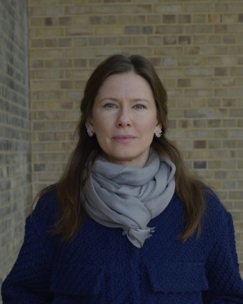 A woman with long brown hair and a gray scarf wears a dark blue coat.