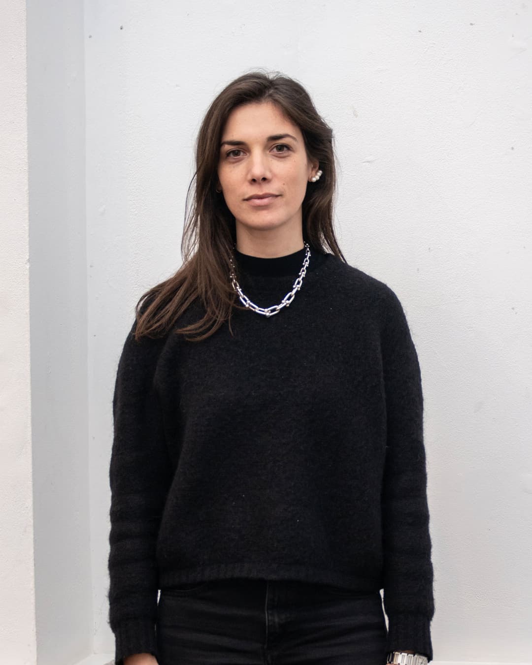 A young woman with long brown hair and dark clothing stands against a white wall.