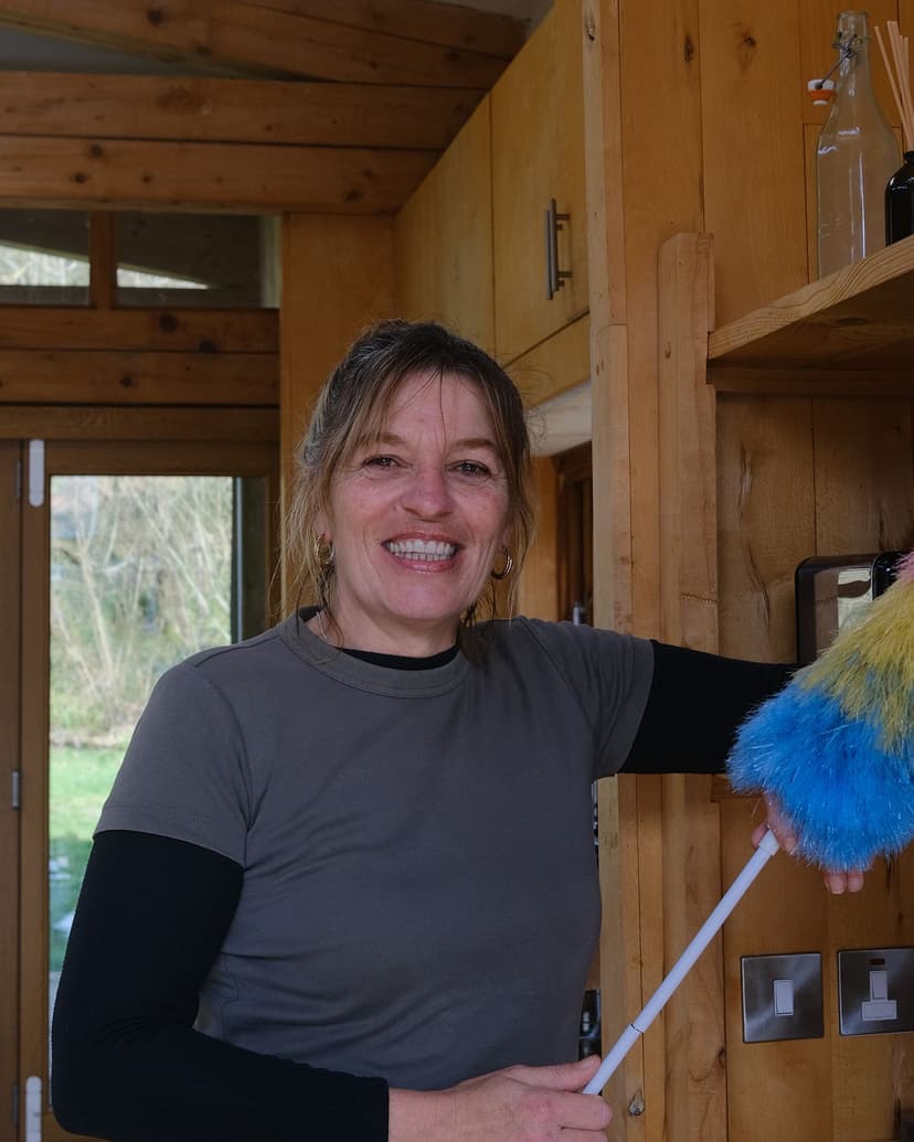 A shite skinned woman with blonde hair stands smiling in a timber clad room, holding a duster.