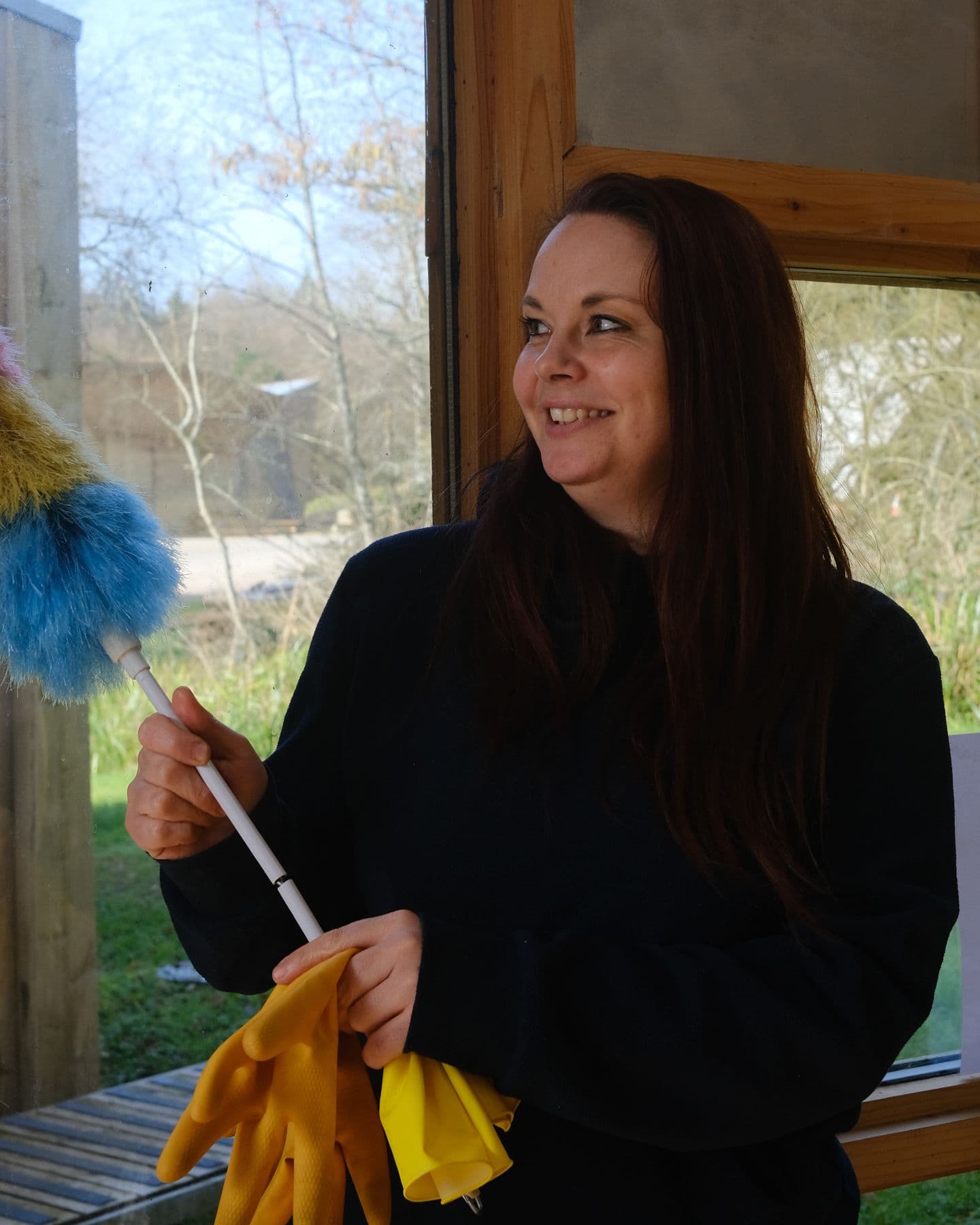 A portrait of a woman with long dark hair holding a duster, against a window looking onto the woodland
