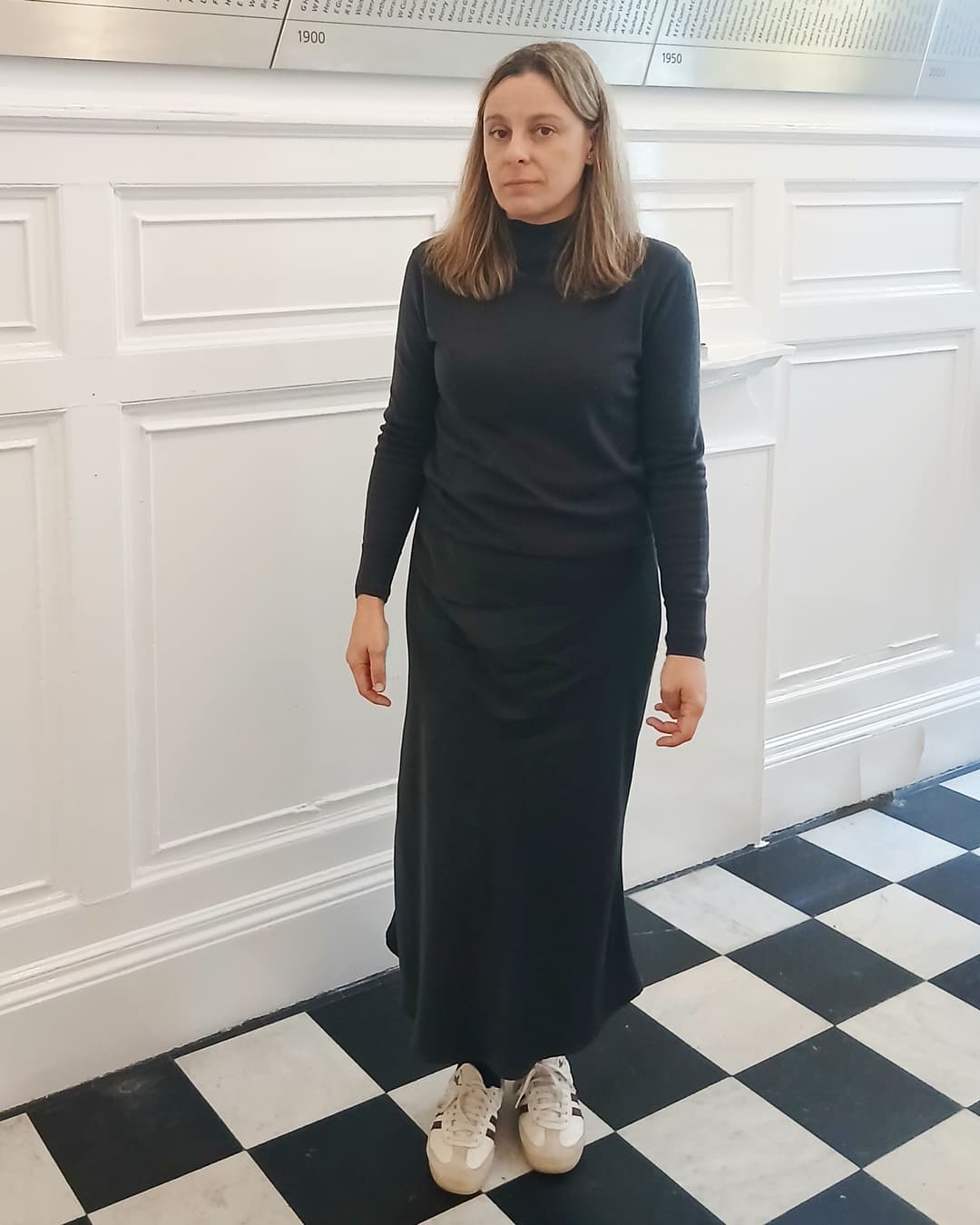 A woman with long brown hair wears a black dress and stands on a black and white tiled floor.