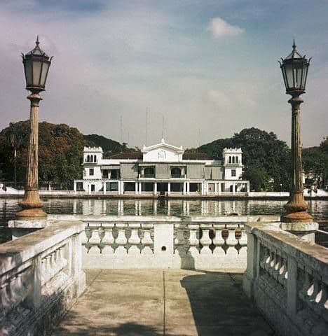 A grand white building with multiple windows is reflected in a calm body of water between two ornate lampposts.