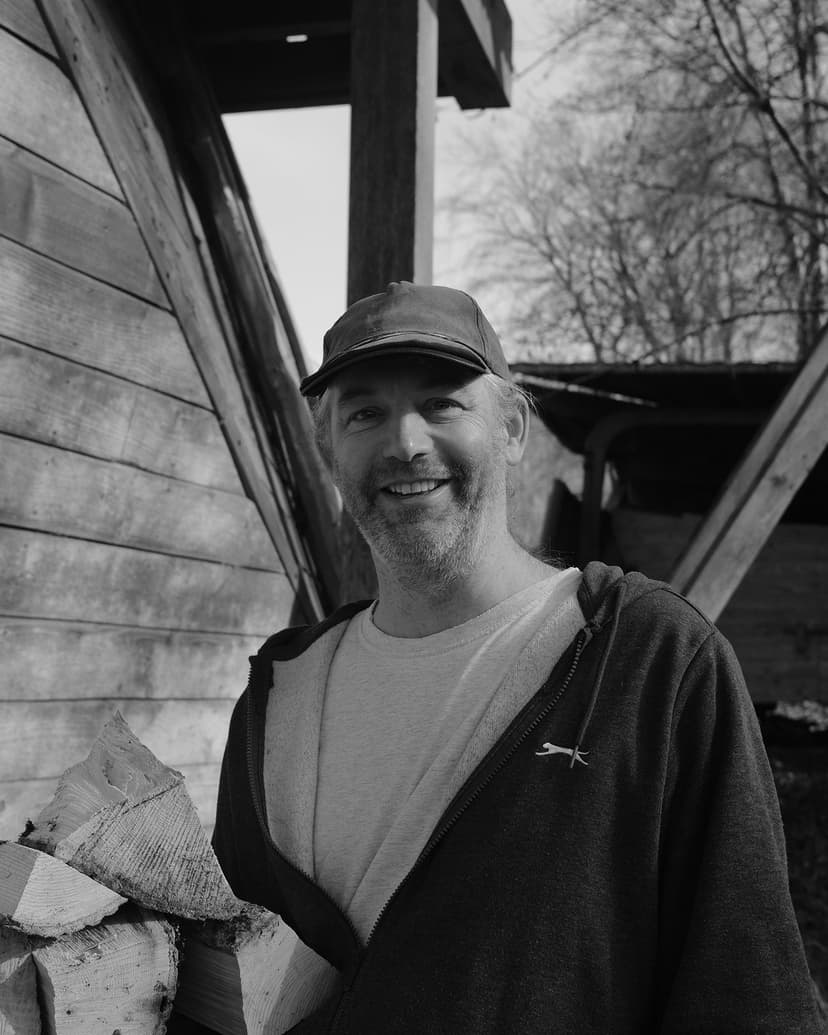 A black and white photo of a white skinned man wearing a baseball cap, standing in front of a timber building and trees.