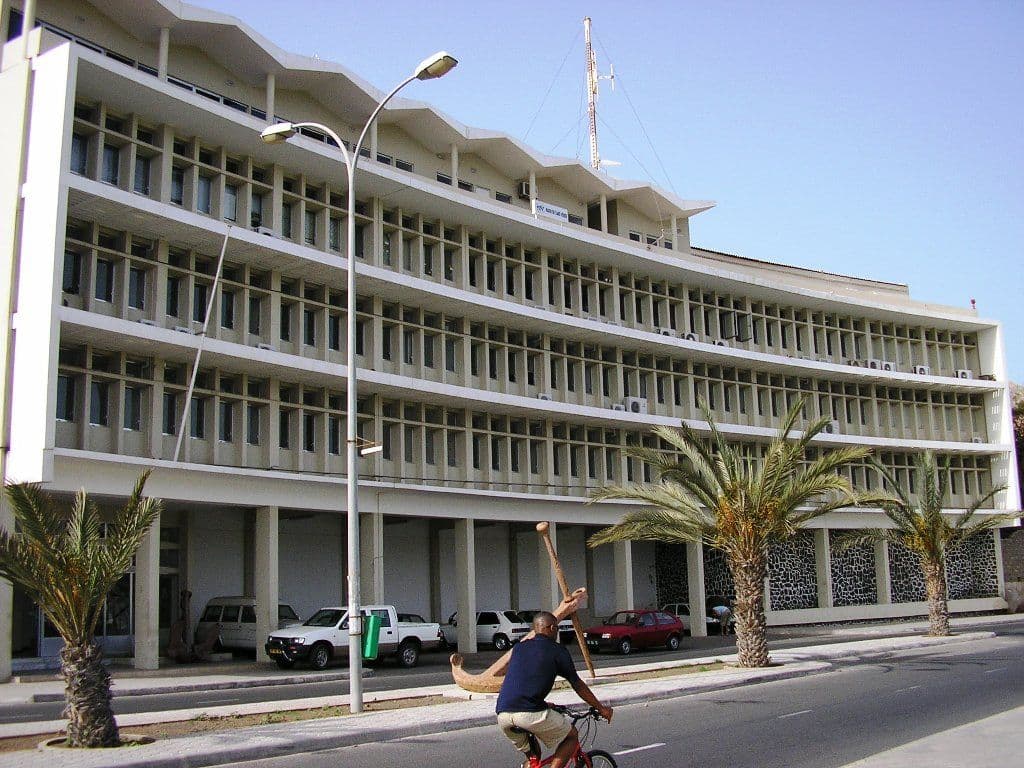 A large, multi-story white building with many windows and palm trees lining the street.