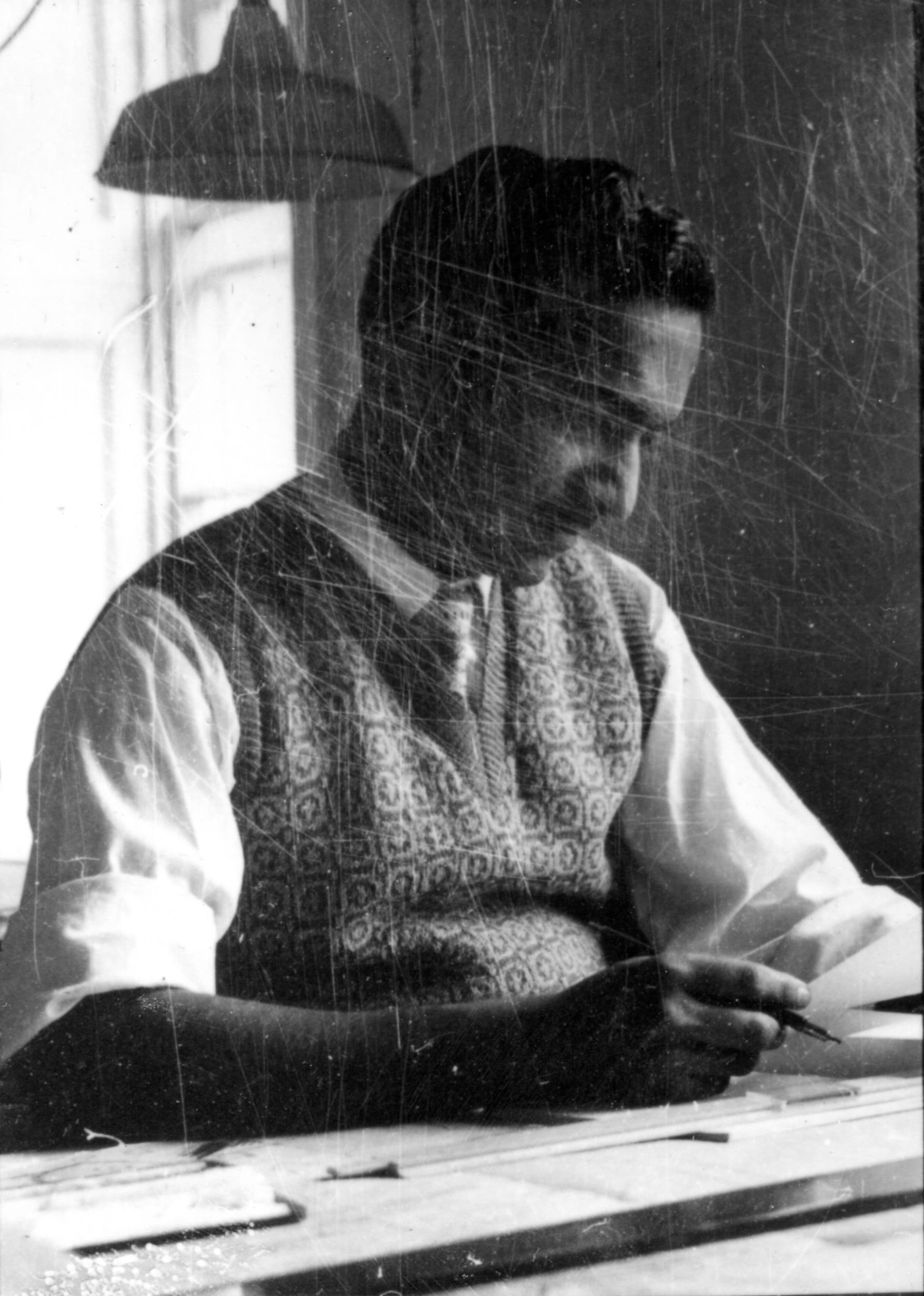 A man in a vest and collared shirt is sitting at a desk, drawing with a pencil.