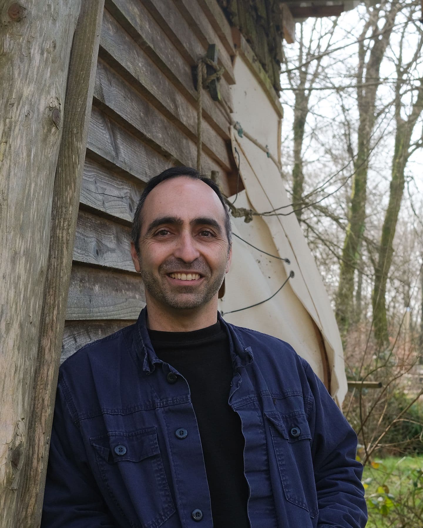 A white skinned man with short dark hair stands against a timber building in a woodland.