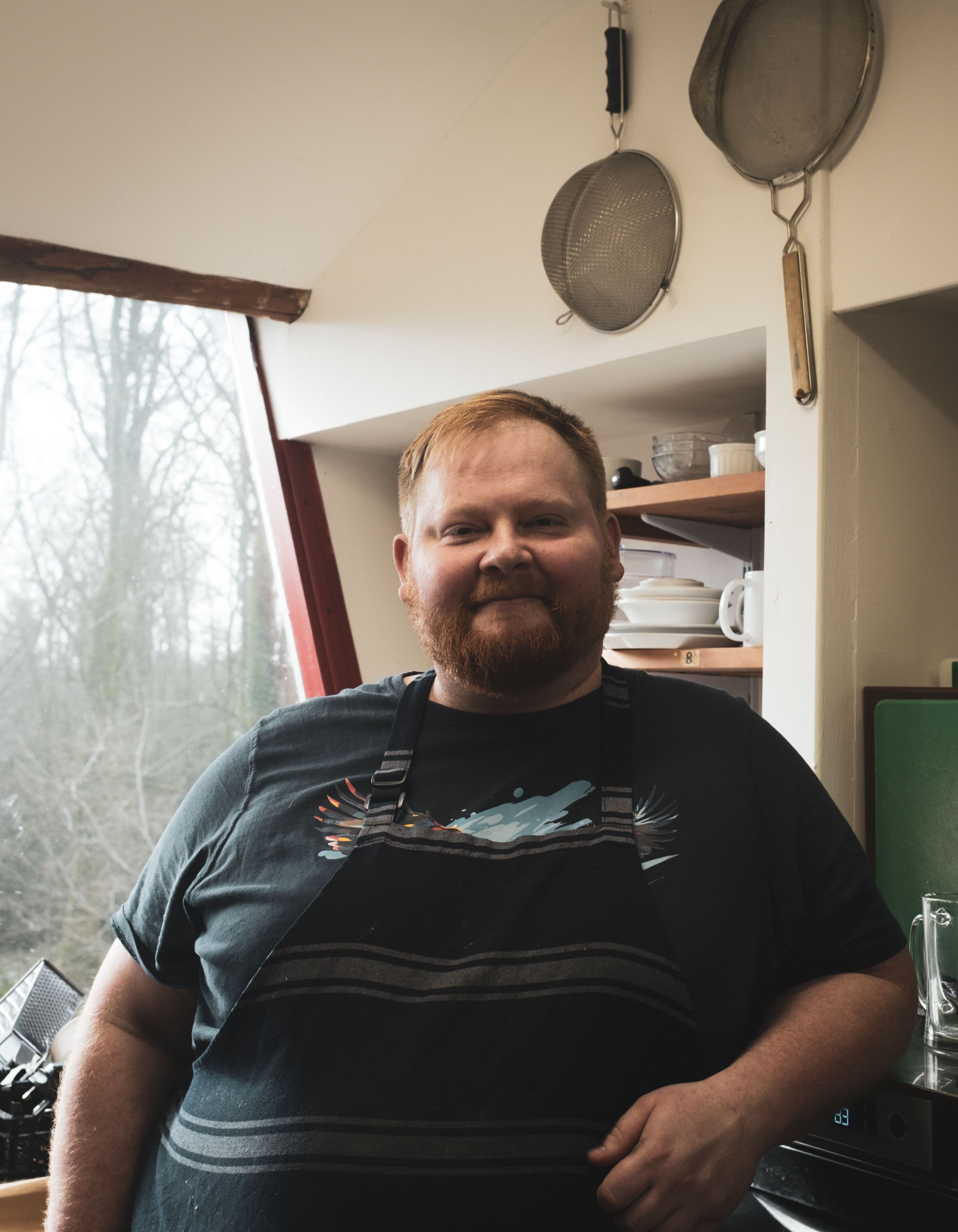 A man with a beard stands in a kitchen in front of a window looking out onto the woodland.