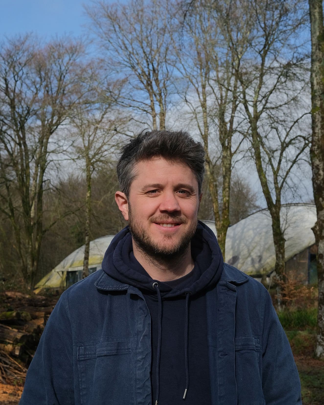 A white skinned man wearing a blue jacket stands in a woodland, with a curved roof visible in the background.