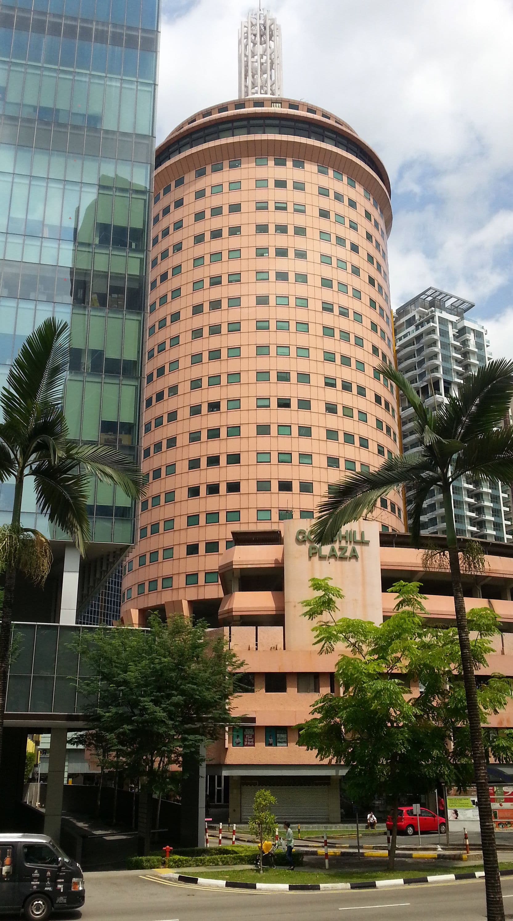 A circular peach-colored building with many windows stands tall amidst modern glass skyscrapers and lush green trees.