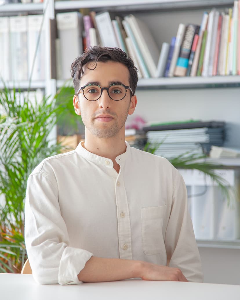 A man with glasses and a white collared shirt sits at a desk in front of a bookshelf.