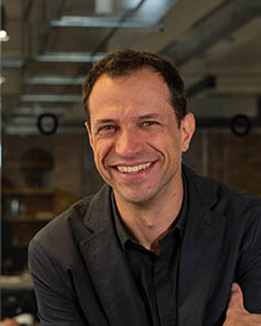 A smiling man with short brown hair, wearing a dark collared shirt and a blazer.