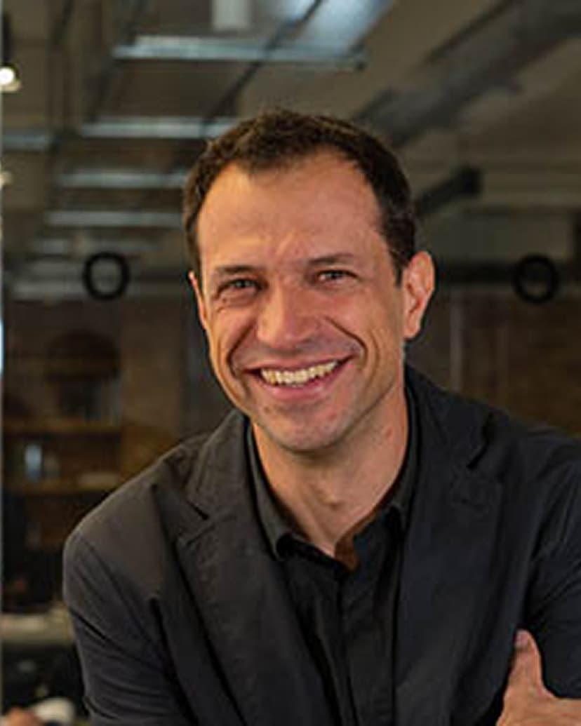 A smiling man with short brown hair, wearing a dark collared shirt and a blazer.
