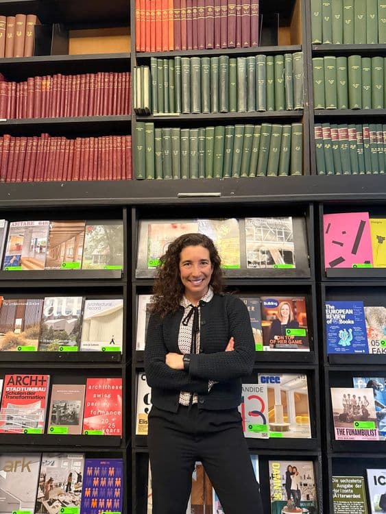 A woman with curly hair smiles, arms crossed, in front of a wall of books.