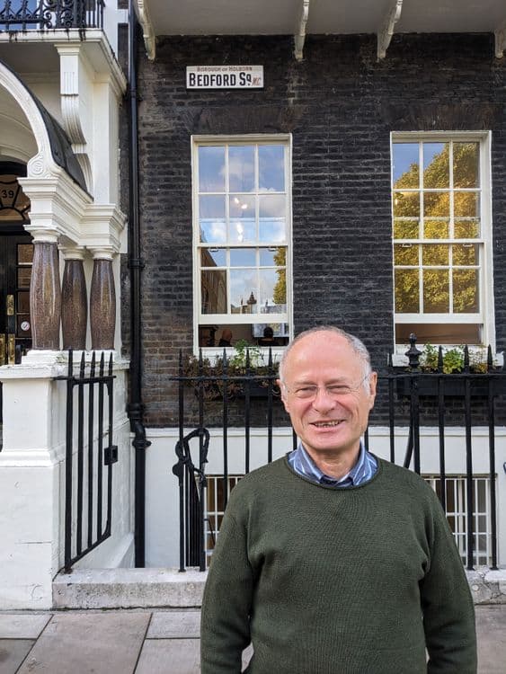 Elderly man with a balding head and a friendly smile stands outside a building.