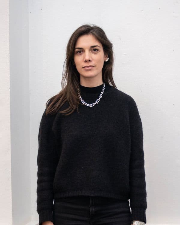 A young woman with long brown hair and dark clothing stands against a white wall.