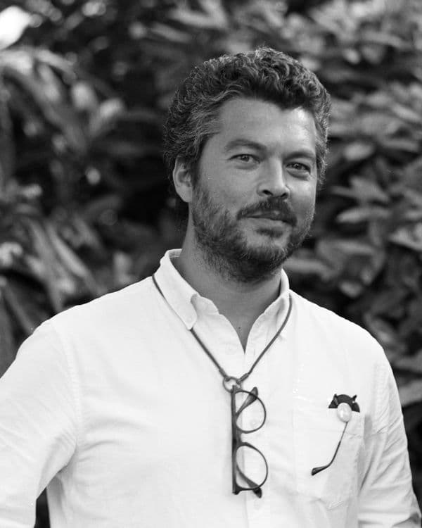 A black and white portrait of a man with short, wavy hair, beard, and glasses.