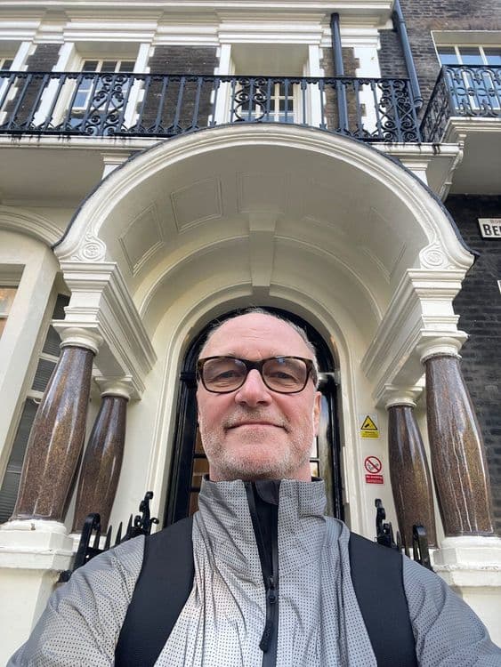 A man with glasses and a gray jacket stands in front of an ornate building entrance.