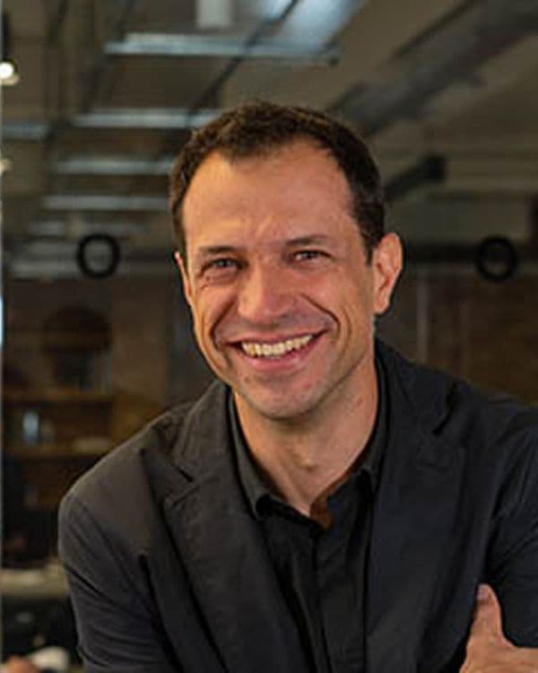 A smiling man with short brown hair, wearing a dark collared shirt and a blazer.