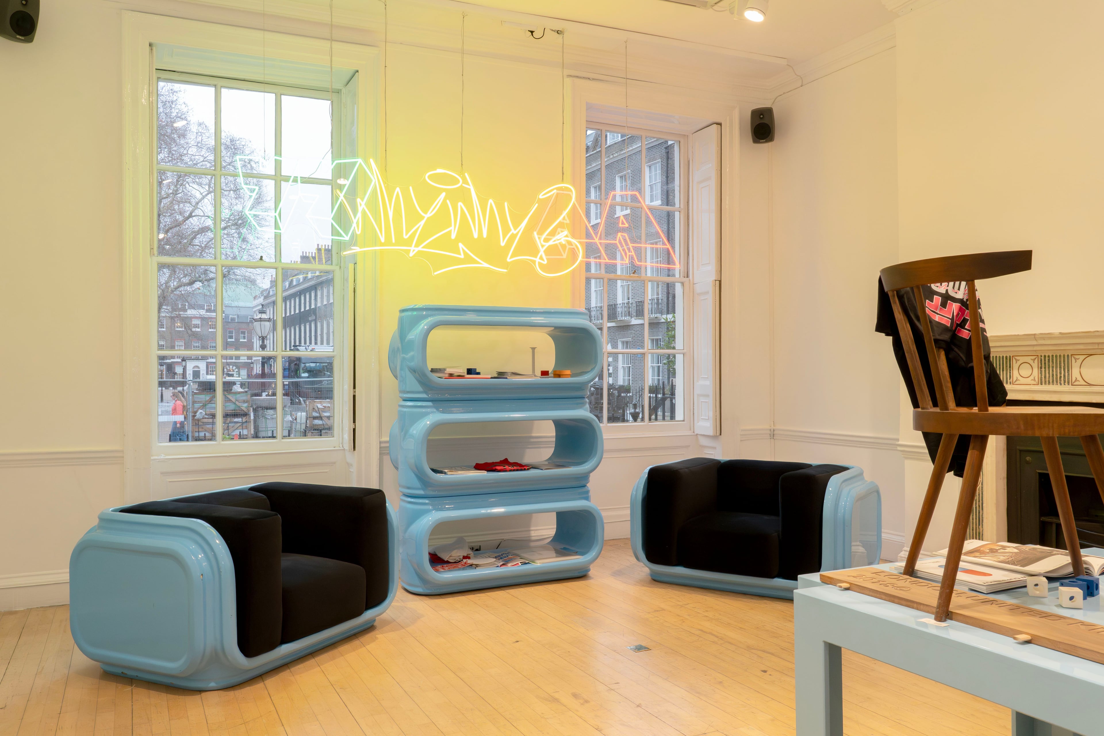 Two blue plastic padded armchairs with black cushions next to a blue plastic bookcase in a gallery in front of windows, with a coloured neon sign above.