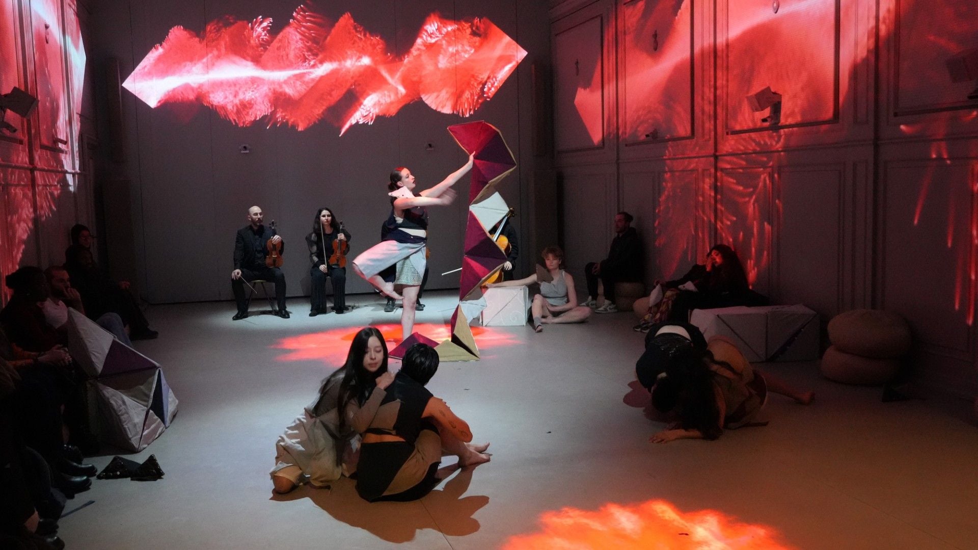 A performance artist with arms outstretched dances under red abstract projections in a dimly lit room.