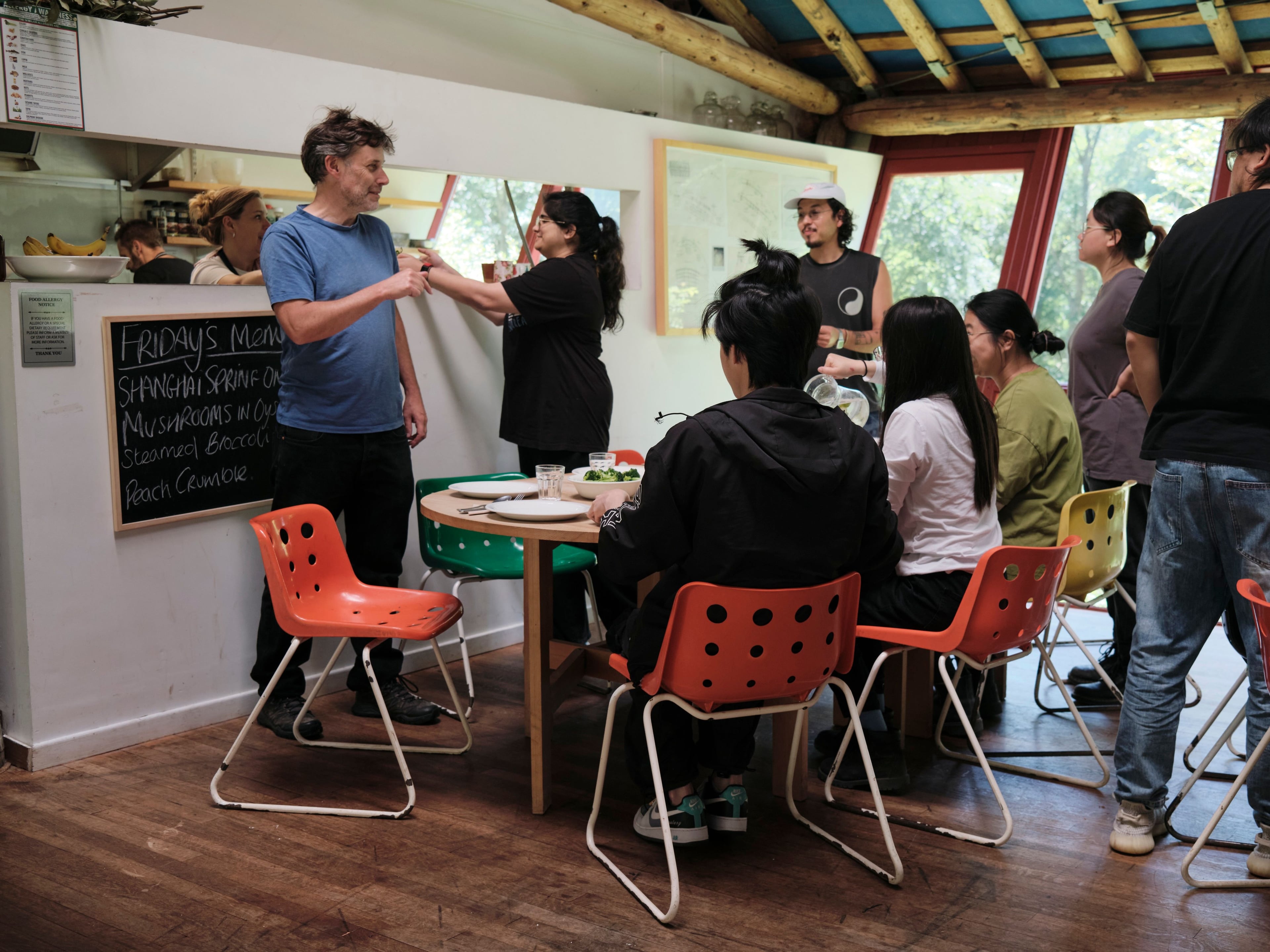 A group of people gathers in a kitchen, watching two women interact at a counter.