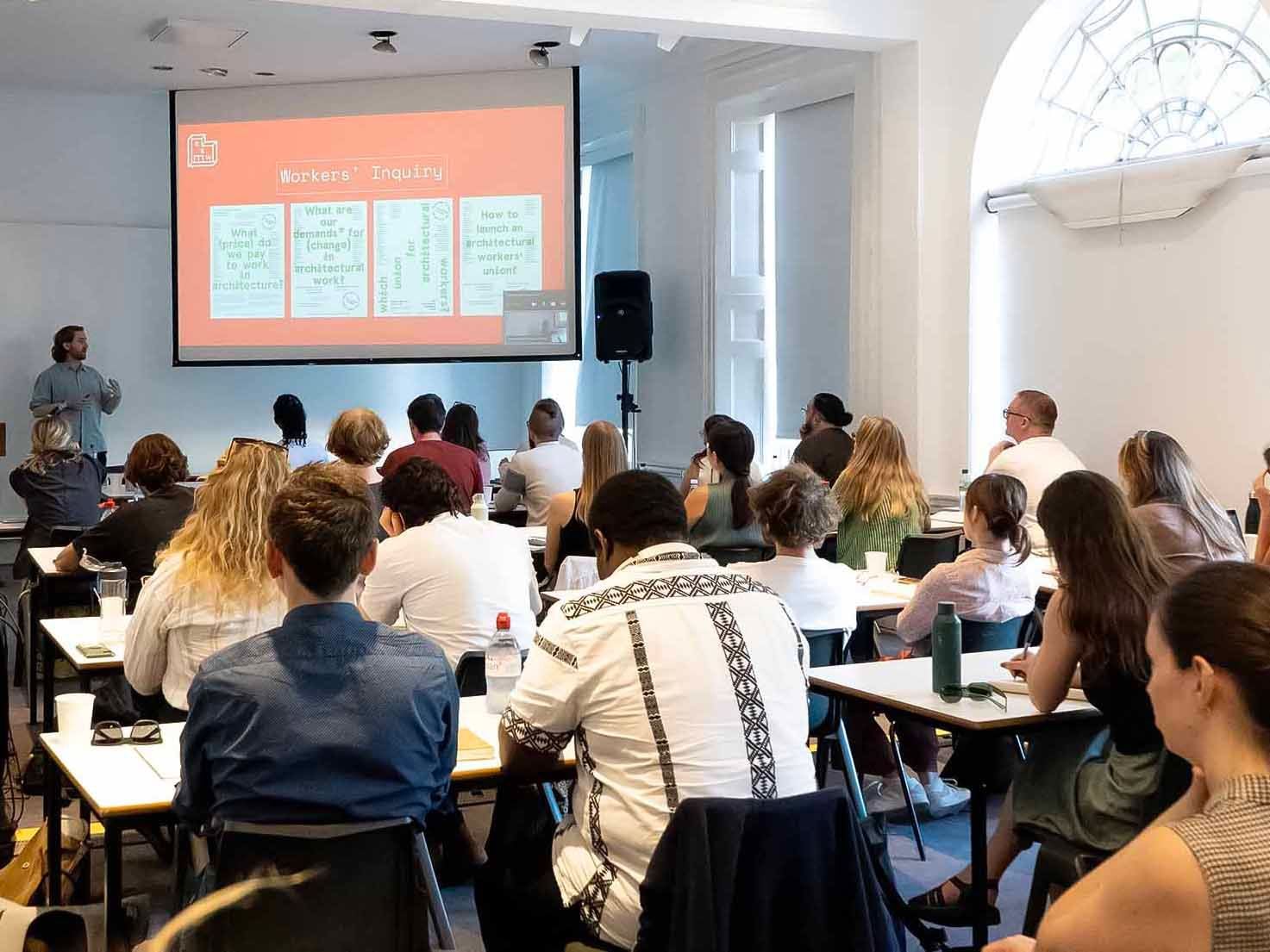 Students sit at desks in front of a speaker and a projector screen in a Georgian lecture hall.
