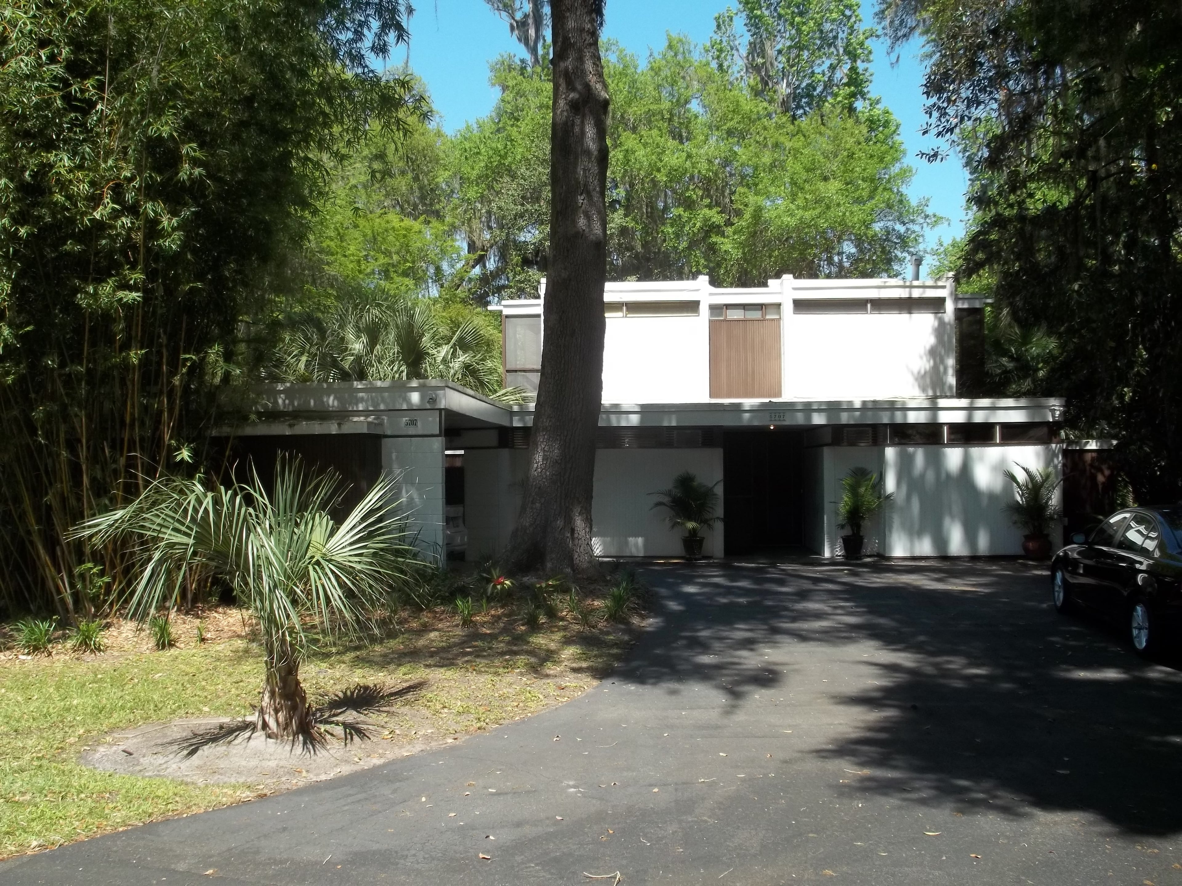 Modern white house with flat roof and geometric windows, surrounded by lush green trees.