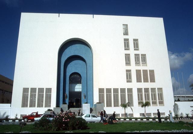 Modern white building with a large blue arched entrance and people outside.