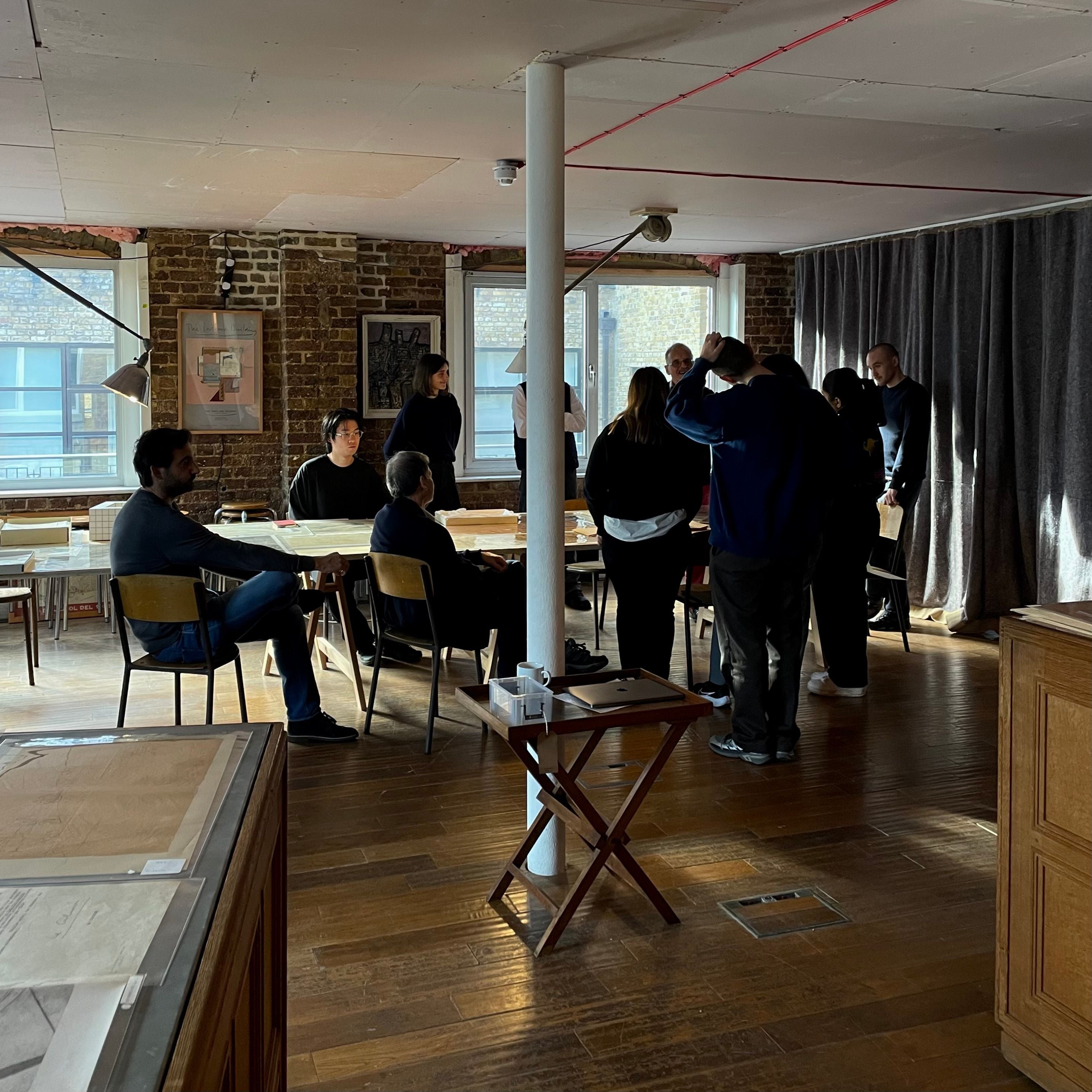 A group of people stand around a table in a brightly lit room with wooden floors.