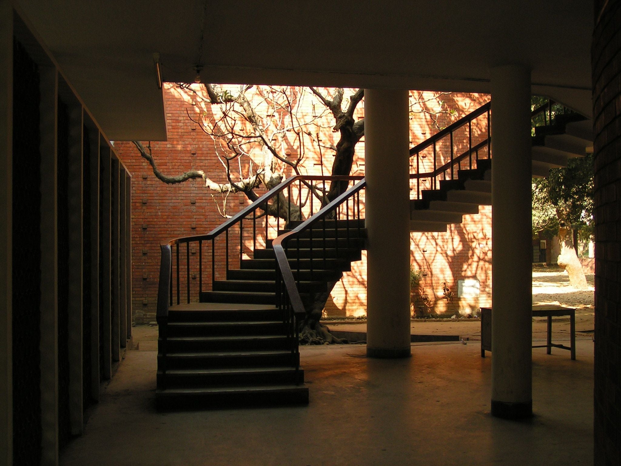 Curving wooden staircase with dark railings ascends towards a sunlit courtyard.