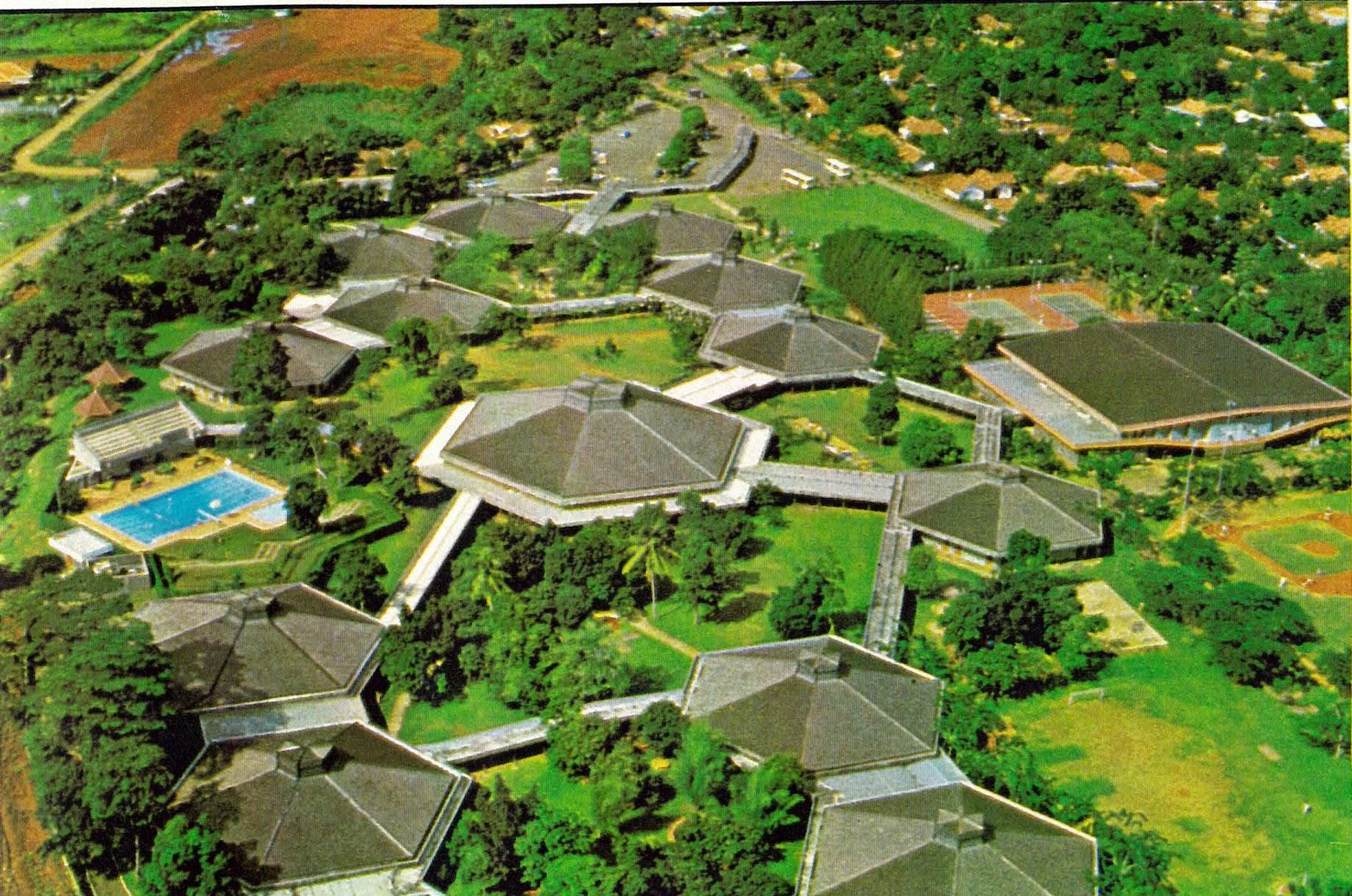 Aerial view of a campus with modern buildings, connecting walkways, swimming pool, and green landscape.