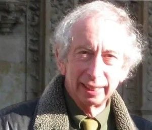A smiling man with white curly hair and a dark jacket looks directly at the viewer.
