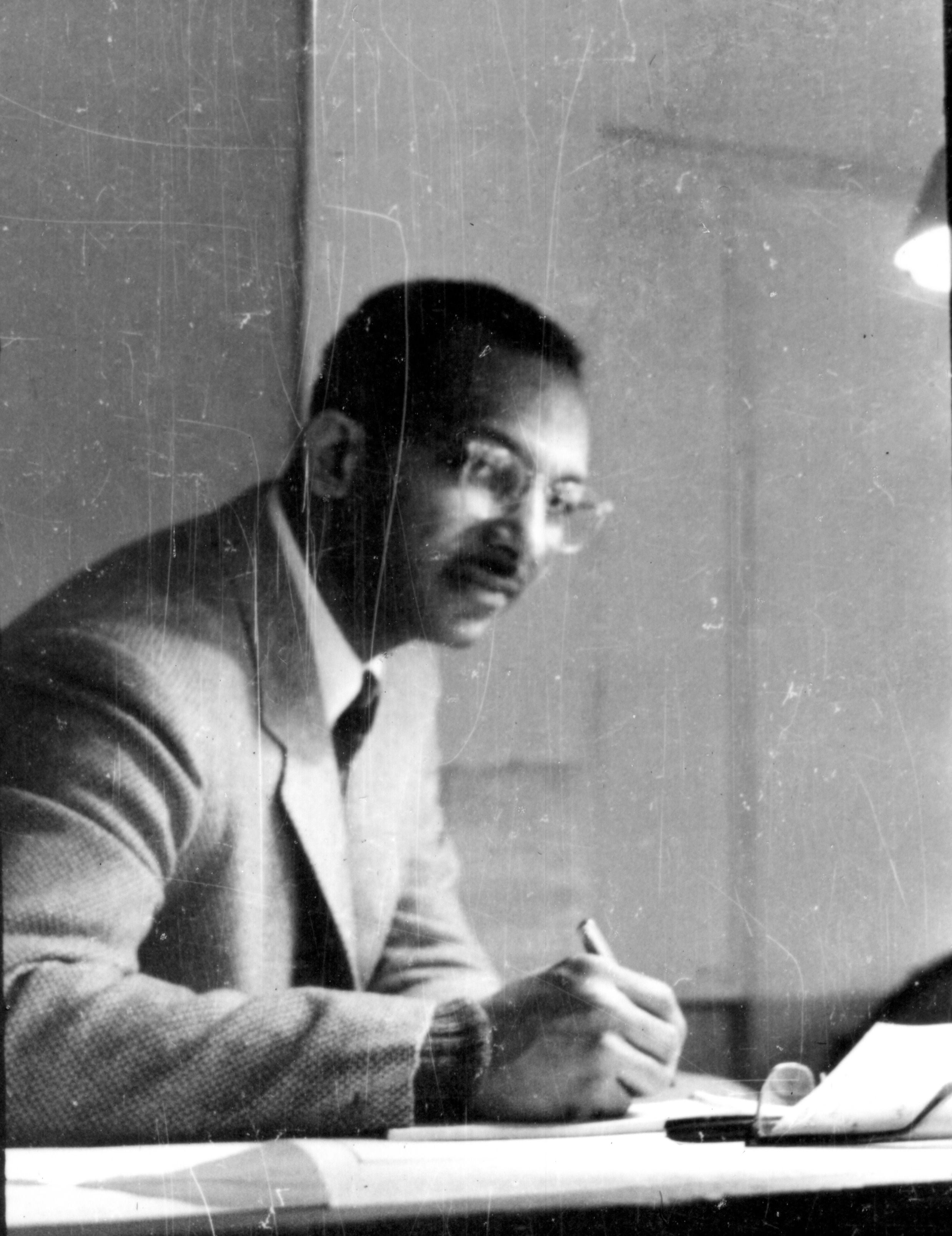 Man in suit and glasses writing at a desk under a lamp.