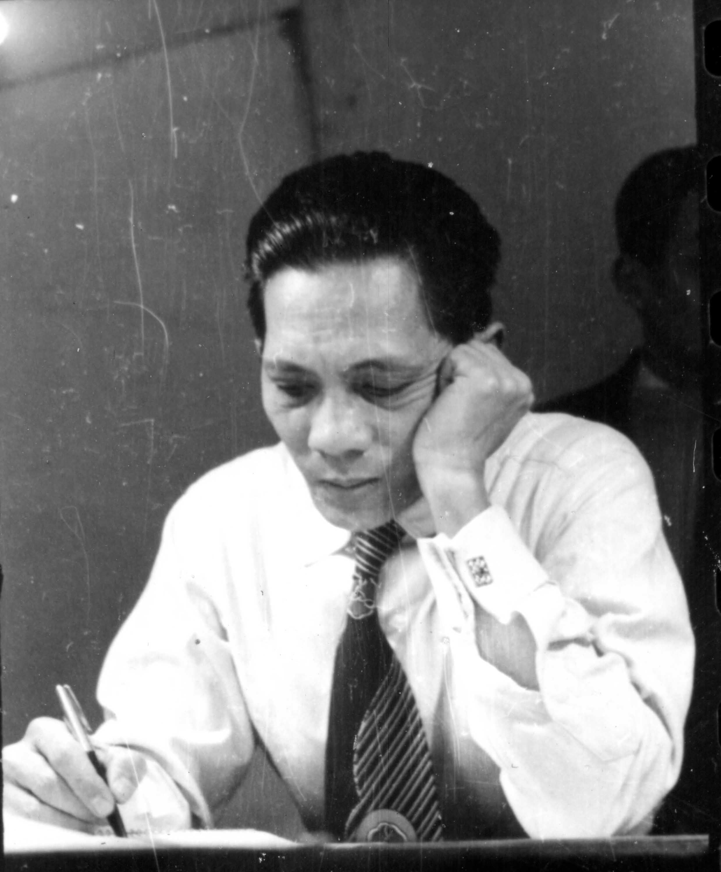 Man in a white shirt and striped tie writing at a desk, head resting on his hand.