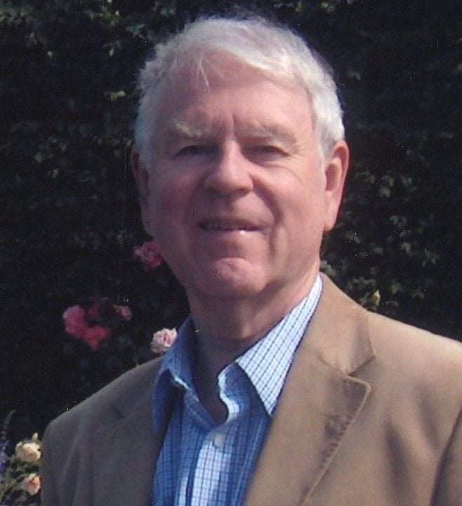 Elderly man with white hair, wearing a blue collared shirt and tan jacket.