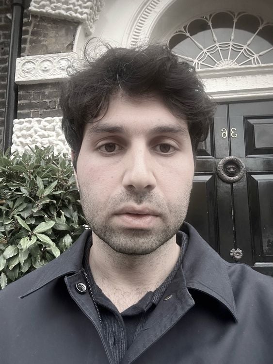 A man with dark, curly hair and a serious expression, wearing a dark collared shirt.