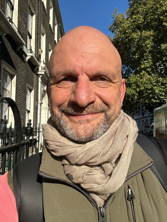 Bald man with a beard smiles, wearing a scarf and jacket outdoors.