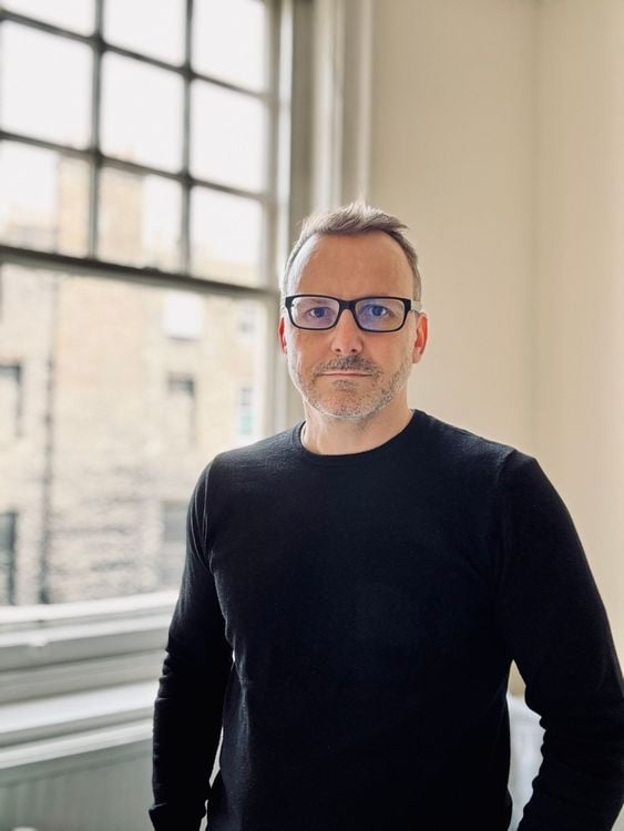 A man wearing glasses and a black sweater stands in front of a large window.
